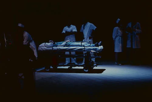 Chris Parnell as Randle P. McMurphy (center) finds himself strapped to the bed as unidentified nurses, orderlies, and doctors stand around him.